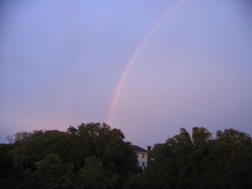 Faint rainbow in dusky sky over dark treeline and a white house gable