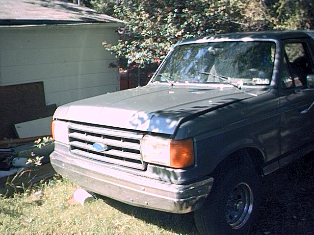 Front 3/4 view of a gray full-size Ford Bronco parked on grass next to a white house