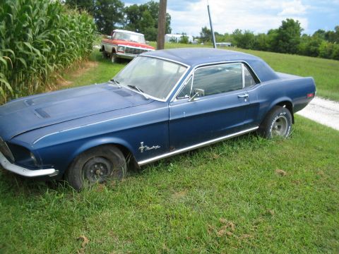 1967 Ford Mustang - A blue 1967 Ford Mustang coupe sits in a grassy field next to a cornfield, with a pickup truck visible behind it.