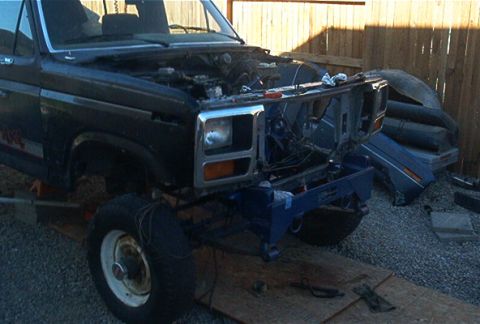 1986 Ford Bronco - This dark blue pickup truck is undergoing front-end work, with the grille and some engine components exposed. The body style suggests a mid…
