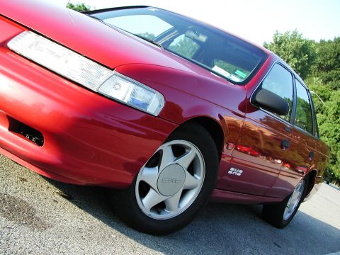 1992 Ford Taurus - Close-up of a red sedan's front quarter panel and wheel, 'SHO' badging on the side.