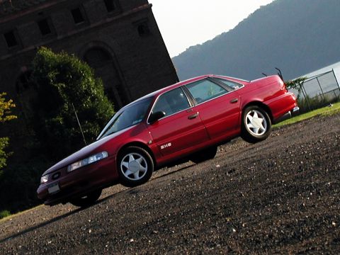 1992 Ford Taurus - Red 1992 Ford Taurus sedan parked on gravel, with a brick building and hills in the background.