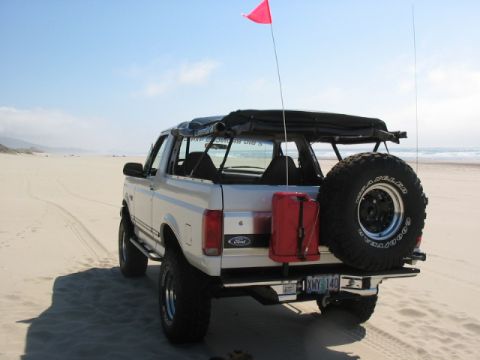 1995 Ford Bronco - Rear view of a 1995 Ford Bronco on a sandy beach, equipped with a spare tire and a red jerry can.