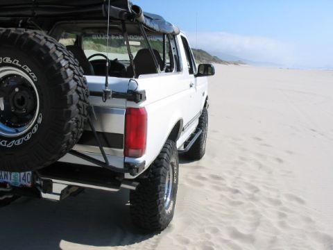1995 Ford Bronco - A white Ford Bronco is parked on a sandy beach, with the ocean and dunes visible in the distance.