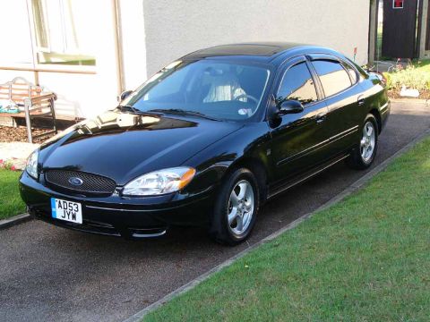 2004 Ford Taurus - A black 2004 Ford Taurus is parked on a paved driveway next to a grassy lawn.