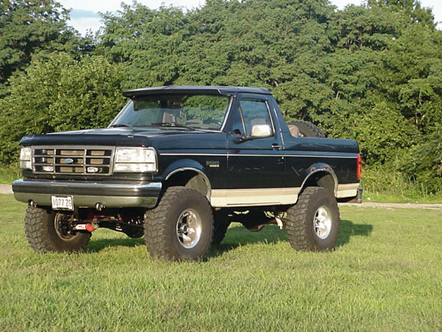 Lifted black 1993 Ford Bronco with off-road tires parked on grass with trees in background