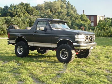 1993 Ford Bronco - This lifted Ford Bronco is parked on a grassy field with trees and a building in the distance.