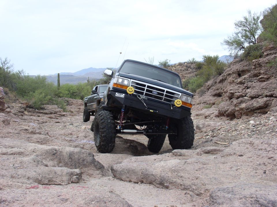 Black lifted Ford Bronco with smiley face decals on bumper, navigating rocky terrain