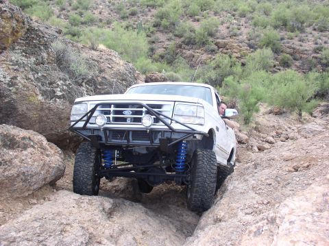 1993 Ford Bronco - A white 1993 Ford Bronco navigates a rocky trail with its front end lifted and blue suspension components visible.