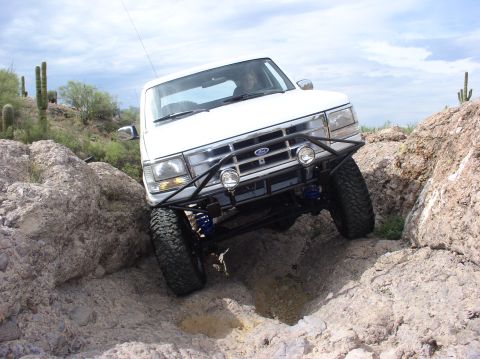 1993 Ford Bronco - White Ford Bronco is shown off-roading over large rocks in a desert landscape.