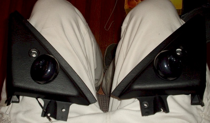Two black triangular A-pillar trim panels with round speakers resting on a lap