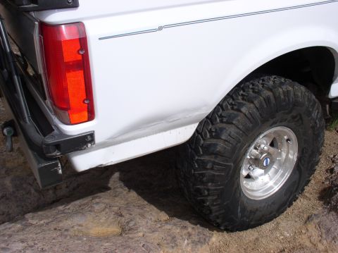 1993 Ford Bronco - Rear view of a white 1993 Ford Bronco showing a dented quarter panel and a large off-road tire.
