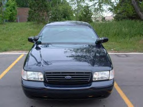 Dark Ford Crown Victoria sedan head-on front view, Ford oval in grille, parked on lot near grass