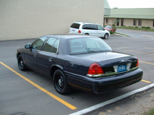 Dark Ford Crown Victoria sedan rear-quarter view in parking lot, minivan and building behind