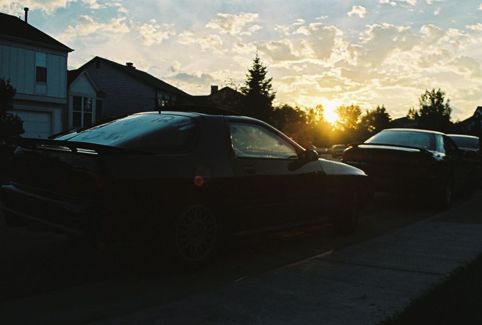 Dark Mazda RX-7 coupe parked on a suburban street, silhouetted against a bright sunset sky