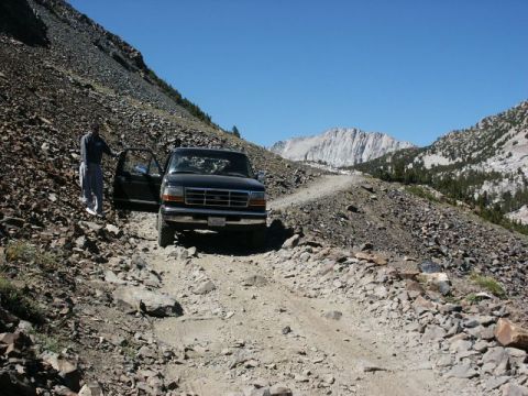 1996 Ford Bronco - A person stands next to a black Ford truck on a rough, unpaved mountain road with steep, rocky slopes on either side.
