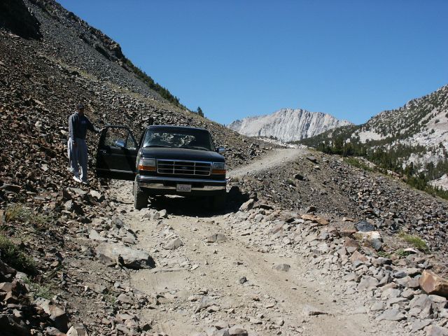 Black Ford truck with driver's door open on a steep, rocky mountain road. Mountains and clear blue sky in background