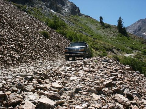 1996 Ford Bronco - A dark SUV navigates a treacherous, rocky mountain path under a clear blue sky.