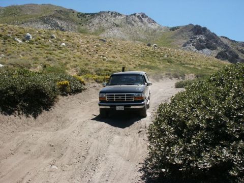 1996 Ford Bronco - A dark Ford F-Series truck navigates a dusty mountain road under a clear blue sky.