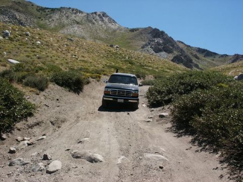 1996 Ford Bronco - A dark SUV navigates a rough dirt road through a mountainous, arid landscape under a clear blue sky.