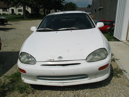 White Mazda MX-3 front view, showing faded paint and dirt. A red car is visible in the background