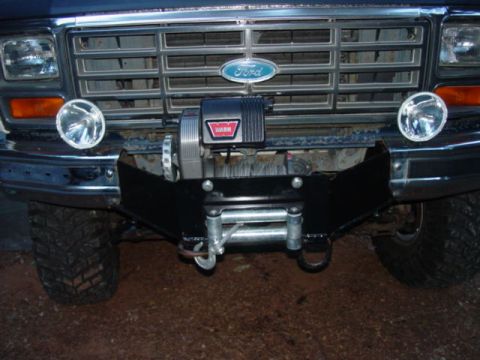 1986 Ford Bronco - Front-end close-up of a blue Ford truck showing a Warn winch with roller fairlead mounted on a black steel plate behind the chrome bumper,…