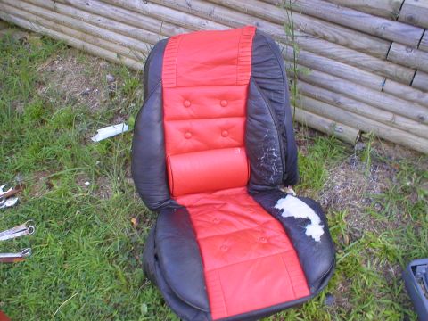 1986 Ford Bronco - A red and black car seat is shown outdoors, resting on grass in front of a wooden fence. The seat exhibits signs of damage and wear.