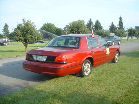 1999 Ford Crown Victoria - This red 1999 Ford Crown Victoria, seen from the rear, appears to be a former police vehicle, complete with a sheriff's star emblem and an…