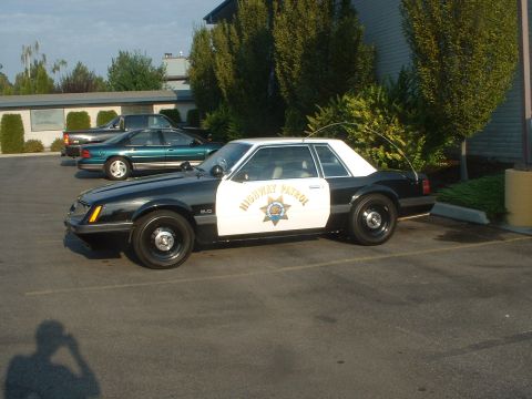 1999 Ford Crown Victoria - A two-tone patrol car with 'HIGHWAY PATROL' text is parked in a lot with other vehicles visible.
