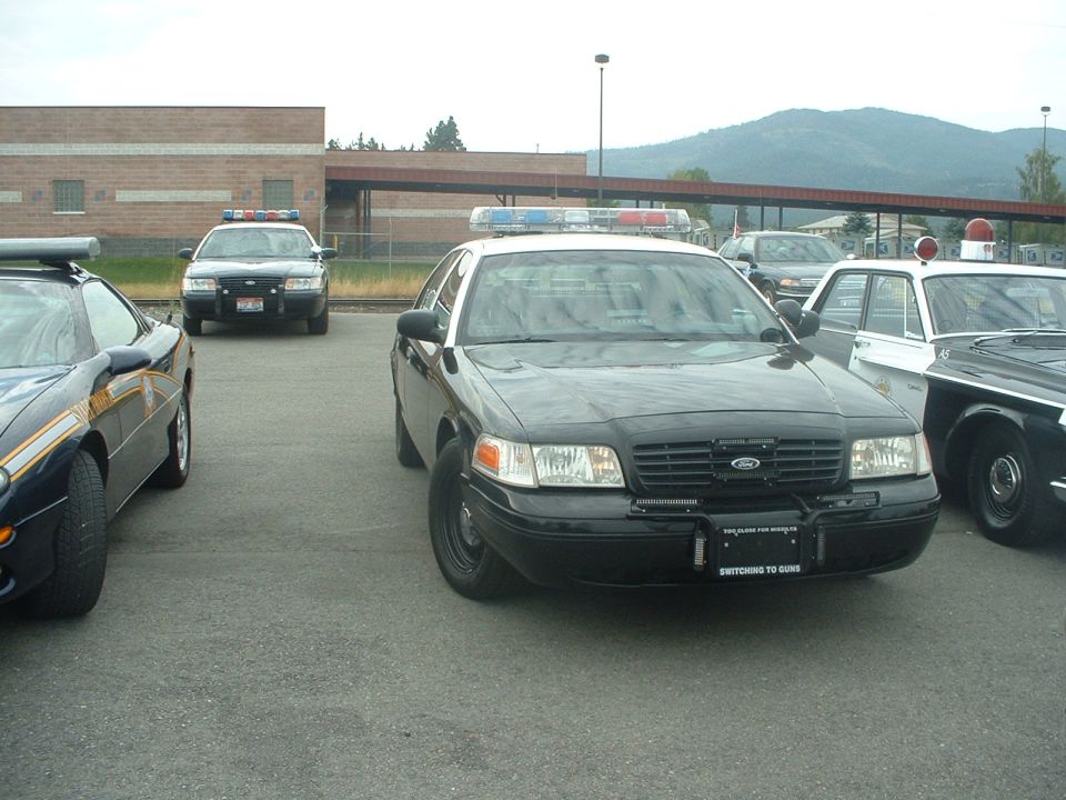 Black police car with light bar, front view. License plate reads 'SWITCHING TO GUNS'