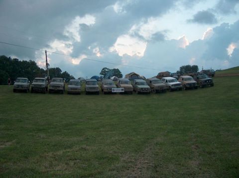 1994 Ford Bronco - A collection of SUVs and trucks are parked in a line on a grassy field, with tents visible in the distance under a cloudy sky.