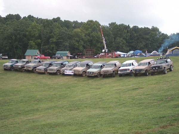 Dozen mud-covered lifted Ford Broncos parked in a row at an off-road event field