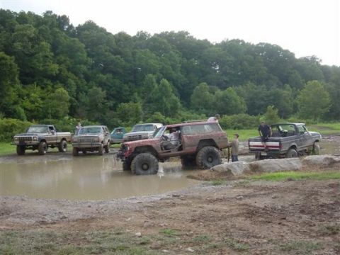 1994 Ford Bronco - Several off-road vehicles are gathered in a muddy area, with one red SUV being assisted by a black pickup truck.