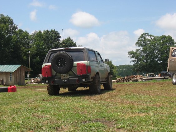 Green Ford Bronco parked in a grassy field, rear view, with spare tire and two red jerry cans mounted on the back