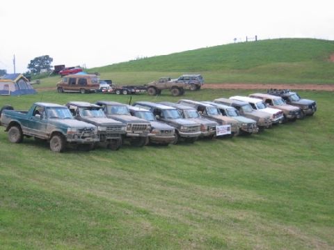 1994 Ford Bronco - A line of trucks and SUVs are parked on a grassy field with a hill in the background.