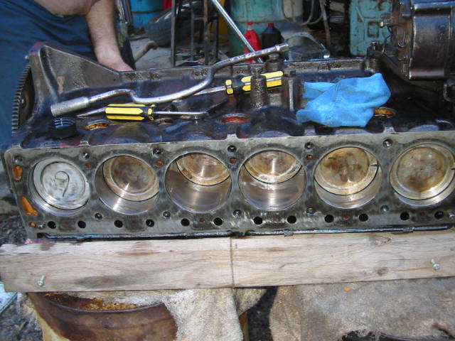 Inline-six engine block with head removed, six pistons visible, tools on top, on a wooden bench.