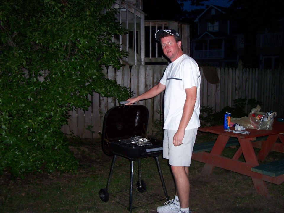 guy in white shirt and shorts by a black charcoal grill in a backyard at dusk