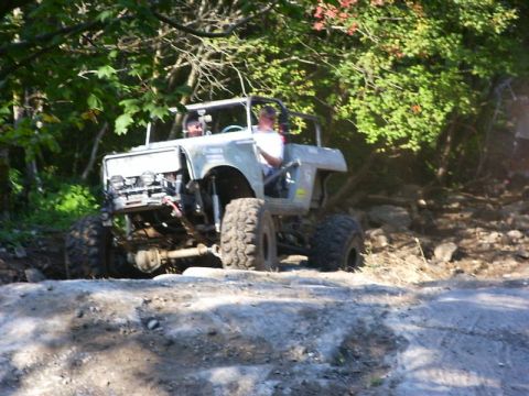 1990 Ford Bronco - A white open-top 4x4 with a roll cage and cut fenders crawls over rocks at the edge of a wooded trail, front tire dropped into a hole. It c…