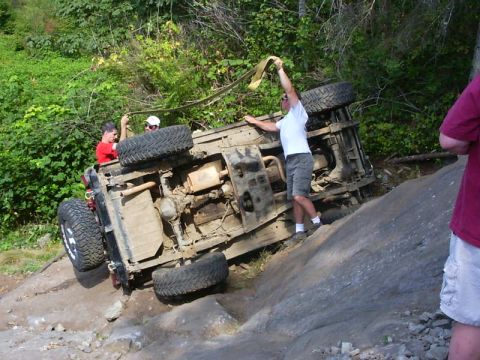 1990 Ford Bronco - A 4x4 lies rolled onto its side on a rock slab with its undercarriage exposed, showing the skid plates, transfer case, driveshaft, muffler…