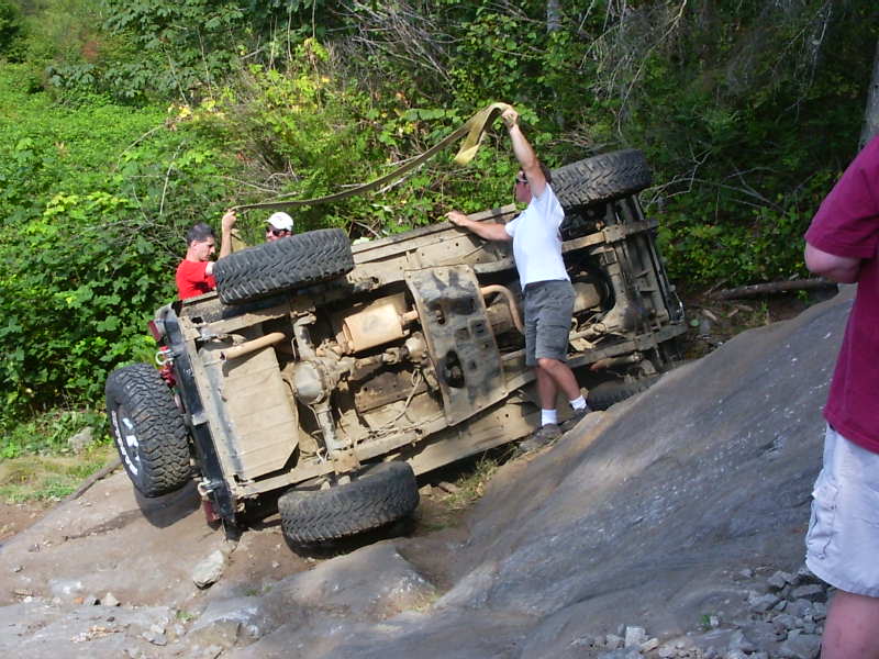 Rolled 4x4 on a rock slab showing undercarriage while men pull a recovery strap