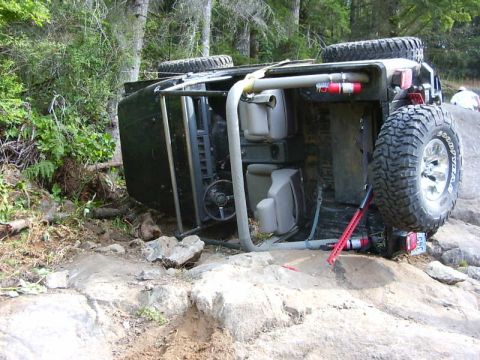 1990 Ford Bronco - A dark 4x4 lies rolled onto its driver's side against a rock ledge on a wooded trail, exposing the open cockpit with grey roll cage, tan se…