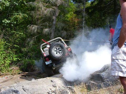 1990 Ford Bronco - Rear view of a caged 4x4 nosed down a rocky bank with a swing-out spare carrier holding a mud tire and a red hi-lift jack strapped to the r…