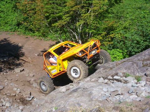 1990 Ford Bronco - A yellow tube-chassis rock buggy with an orange exo-cage climbs a slab of rock on a wooded trail, front tire lifted as it crests the ledge.…