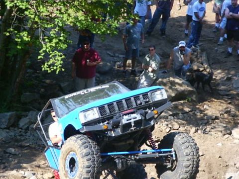 1990 Ford Bronco - A teal Jeep Cherokee-bodied crawler with an exposed roll cage and no doors climbs a rock pile as a crowd of spectators looks on from the ba…