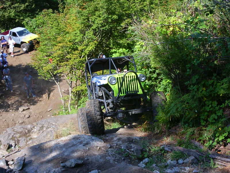 Green off-road buggy navigating a rocky trail with a pickup truck and spectators in the background