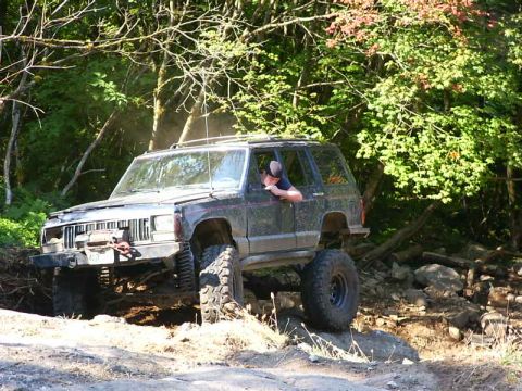 1990 Ford Bronco - A dark-colored Jeep Cherokee XJ navigates a challenging off-road course through a wooded area.