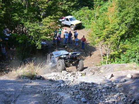1990 Ford Bronco - Elevated view of a caged black buggy on oversize tires working up a rocky quarry climb while a crowd of spectators watches from the trail b…