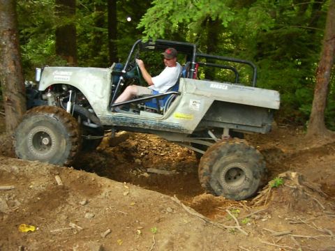 1990 Ford Bronco - A heavily modified off-road vehicle navigates a challenging dirt path surrounded by trees.