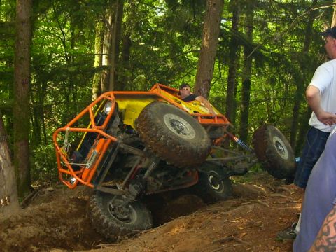 1990 Ford Bronco - A yellow rock buggy in an orange tube exo-cage flexes hard on a rutted dirt climb in the woods, nose pitched up with one front tire drooped…