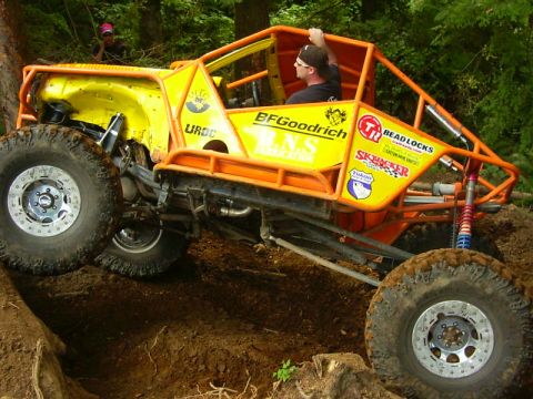 1990 Ford Bronco - Side view of a yellow open-cockpit rock crawler in an orange tube chassis, straddling a dirt trench with the driver looking back over the c…